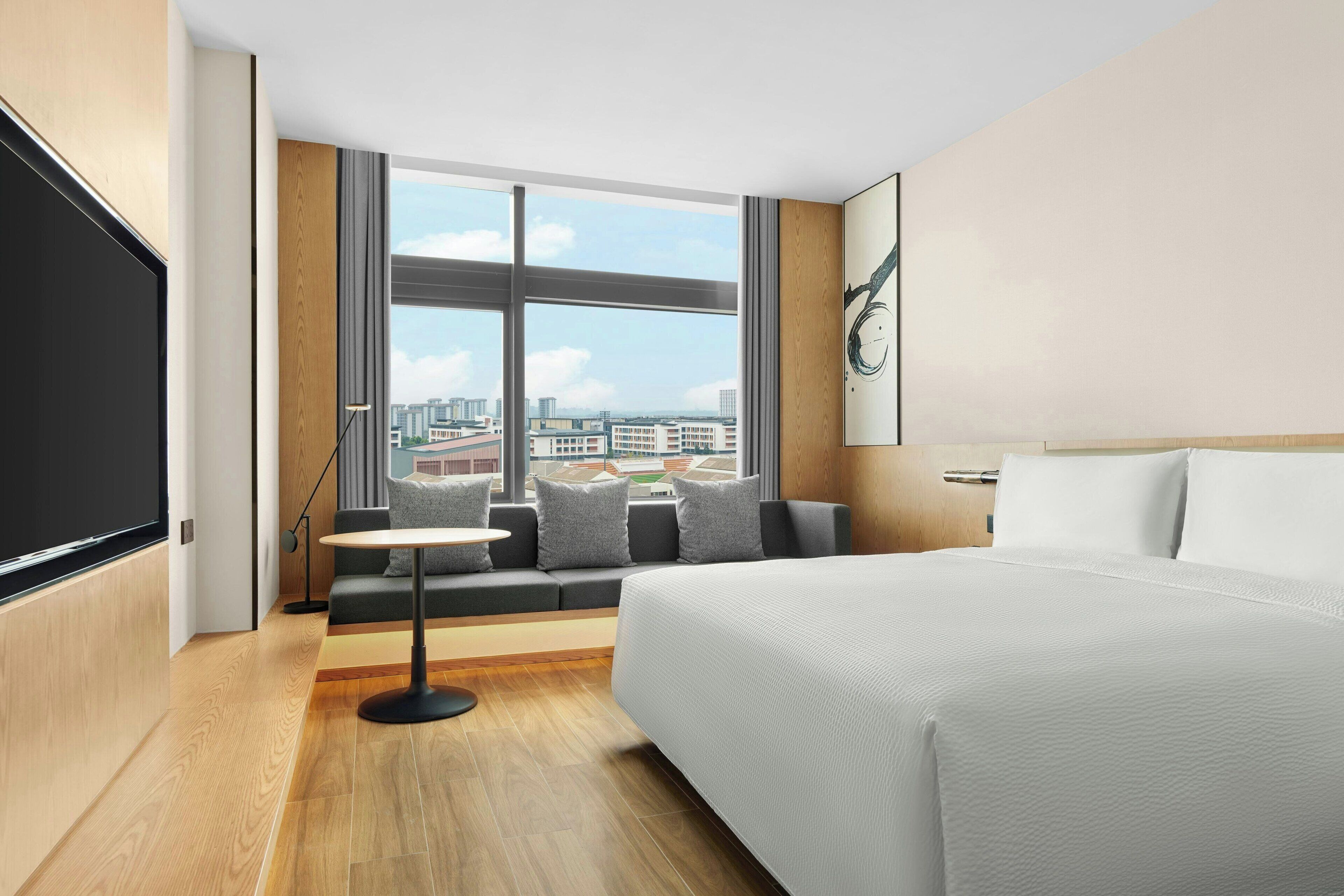 Habitación de hotel moderna con una cama grande, un sofá con cojines, una mesita y un ventanal que ofrece vistas a la ciudad.