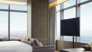 A modern hotel room with a large bed, a flat-screen TV, and a view of the city through large windows.