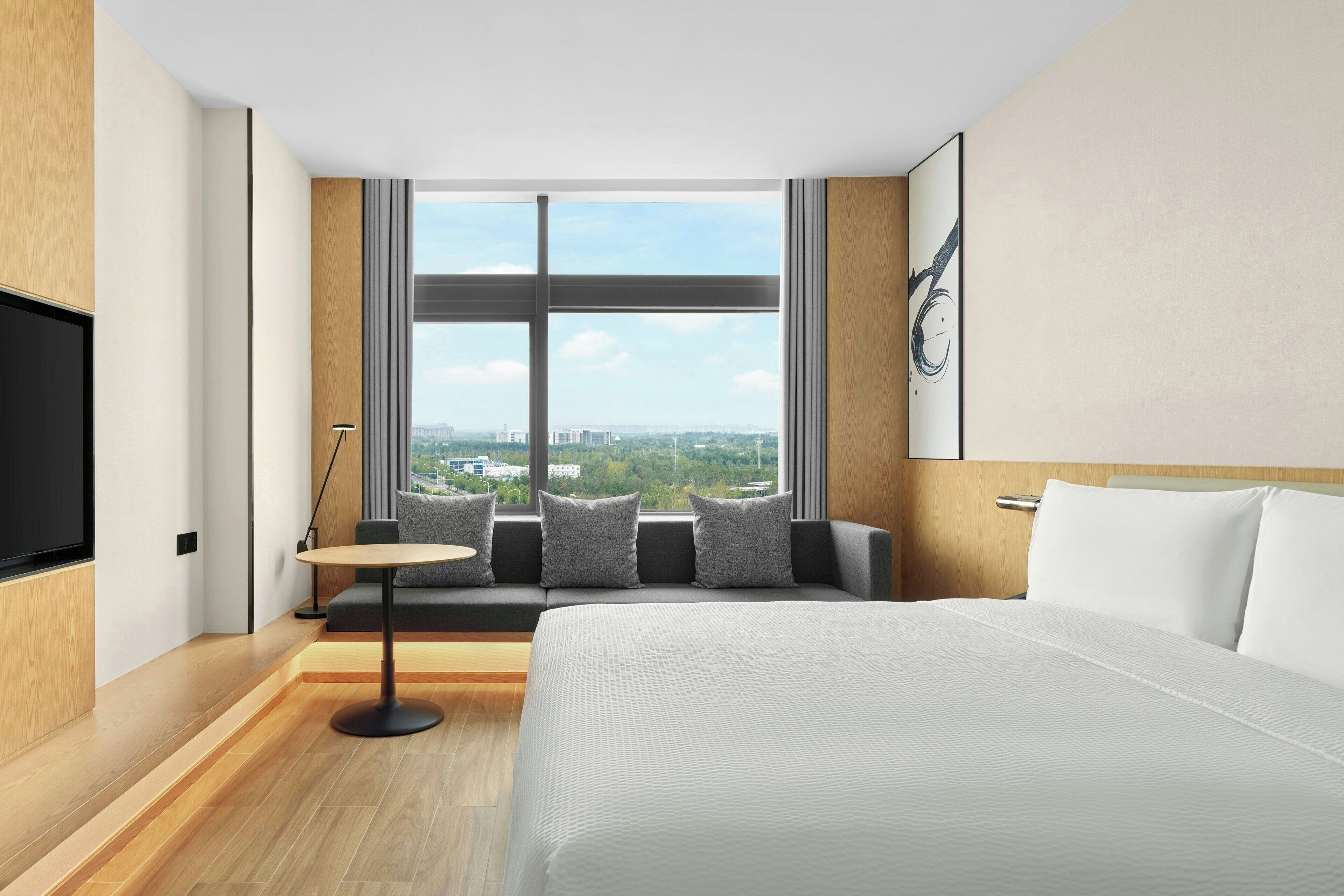 Habitación de hotel con una cama grande, un sofá con cojines, una mesita redonda y un televisor en la pared izquierda.