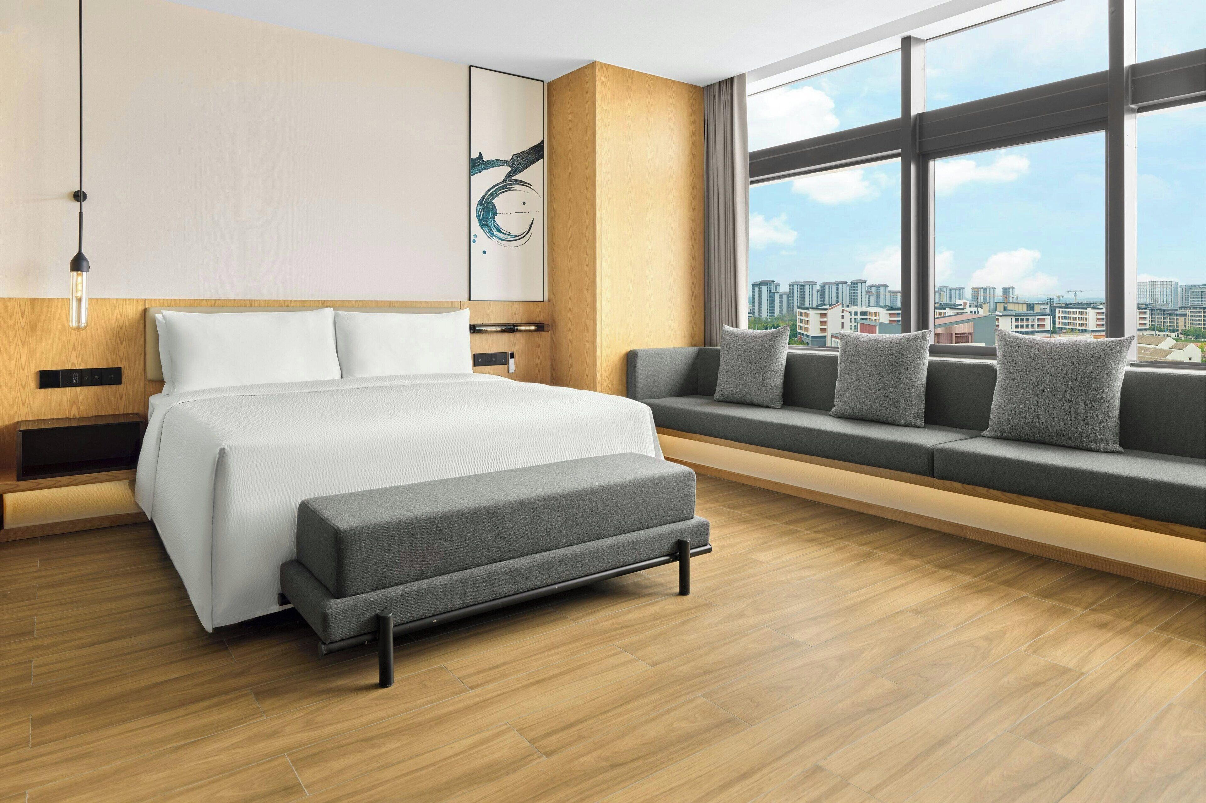 A modern hotel room with a large bed, a sofa with cushions, a window with a city view, and a wooden floor.