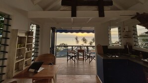 A room with a wooden table, chairs, a bookshelf, and a view of palm trees and a sunset through a large window. - A fusion of classic colonial and cool Mediterranean holiday accommodation (Savusavu)