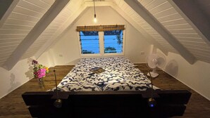 An attic bedroom with a sloped ceiling, a bed with a patterned bedspread, a standing fan, and a window with a view of palm trees. - A fusion of classic colonial and cool Mediterranean holiday accommodation (Savusavu)