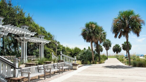 A wooden boardwalk with white railings and benches, surrounded by palm trees and greenery. - Golden Hour Deck Views + Island Fun Await at This Spacious 3BR Retreat (Hilton Head Island)