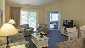 A hotel room with a sofa, two armchairs, a coffee table, a TV, and a balcony with a view of trees. - Golden Hour Deck Views + Island Fun Await at This Spacious 3BR Retreat (Hilton Head Island)