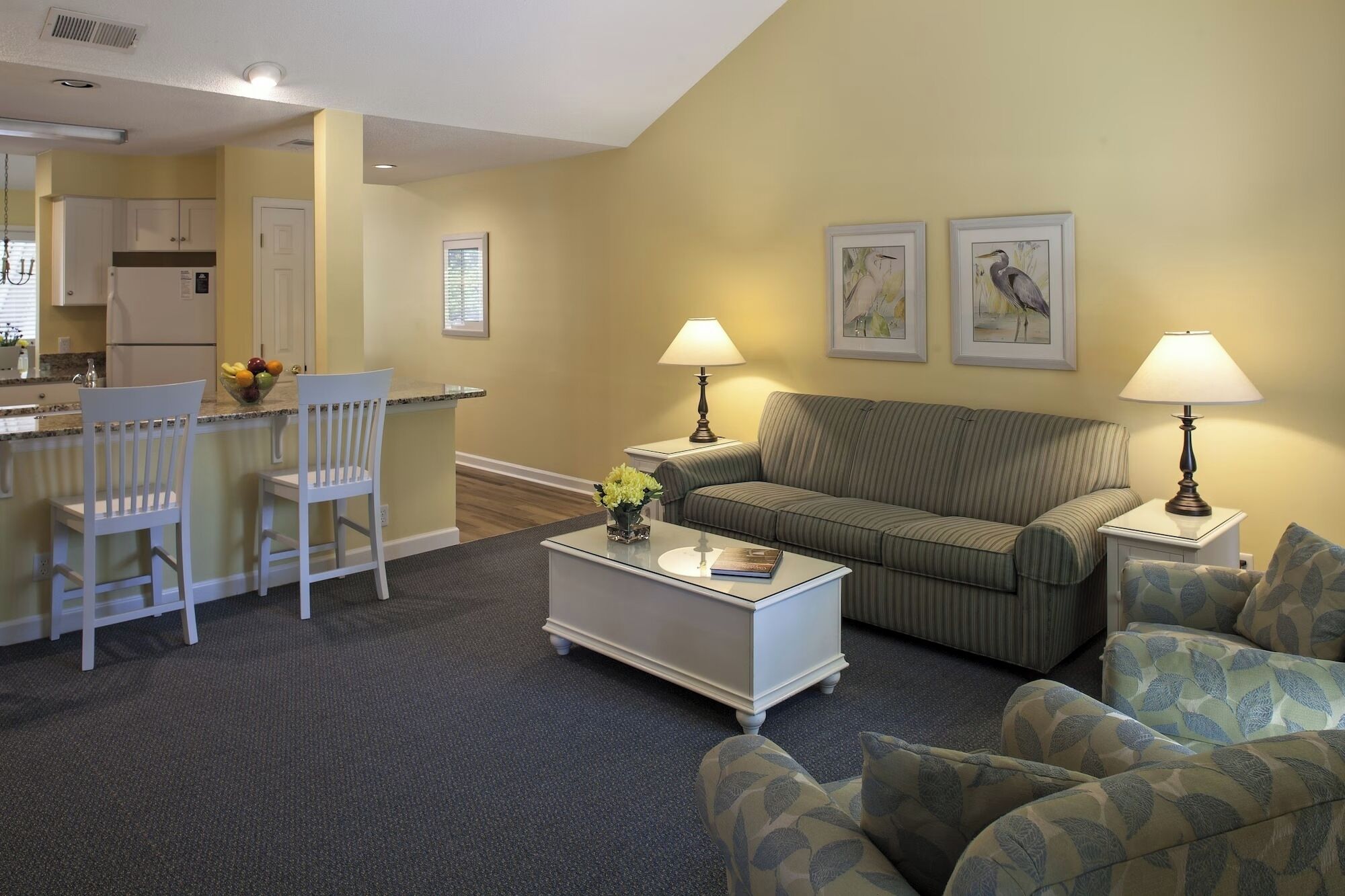 A living room with a sofa, two armchairs, a coffee table, and a kitchen area with a refrigerator and a fruit bowl.