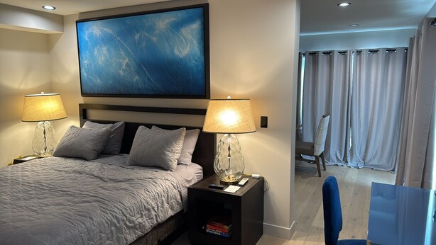 Ein modernes Hotelzimmer mit einem großen Bett, zwei Nachttischlampen, einem Bild über dem Bett und Blick auf einen Balkon mit Vorhängen.