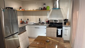 Eine kompakte Küche mit einem Edelstahlkühlschrank, einer Mikrowelle, einem Herd und einem Spülbecken. Darüber befindet sich ein Holzregal und es gibt einen kleinen Esstisch mit Unterlage.