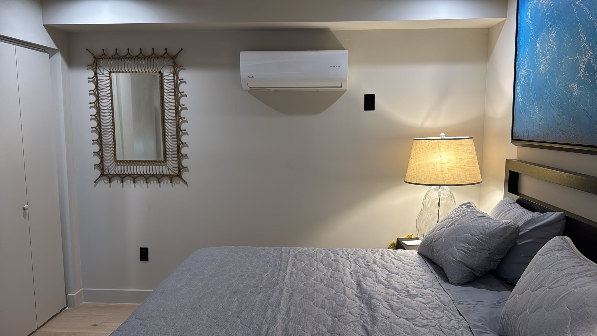 A bedroom with a bed, a wall-mounted air conditioner, a decorative mirror, a bedside lamp, and a painting on the wall.