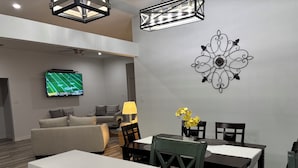 Una sala moderna con un televisor de pantalla plana, un comedor con mesa y sillas, y un elemento decorativo en la pared.