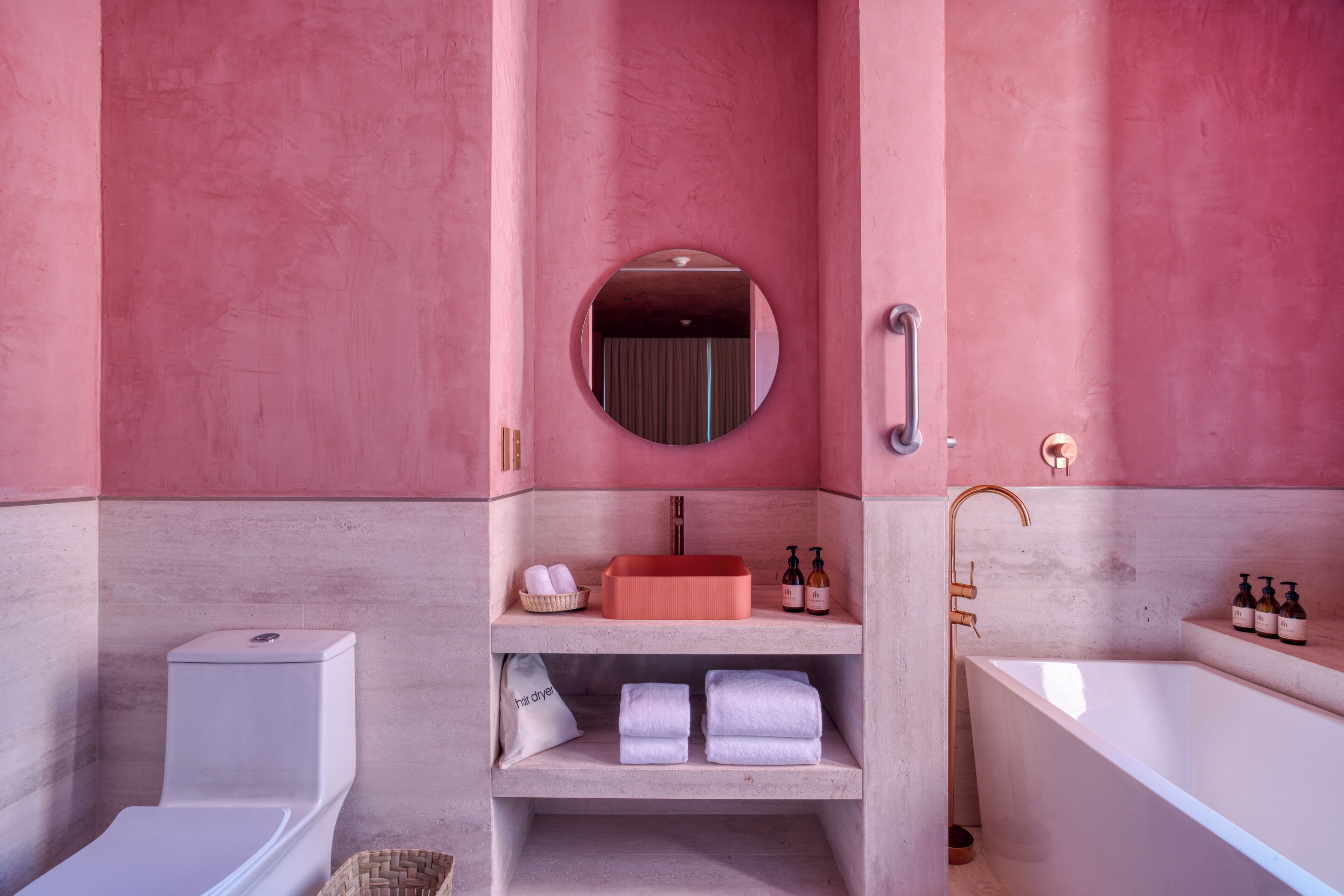 Un baño con pared rosa, espejo redondo, lavamanos, inodoro y bañera.