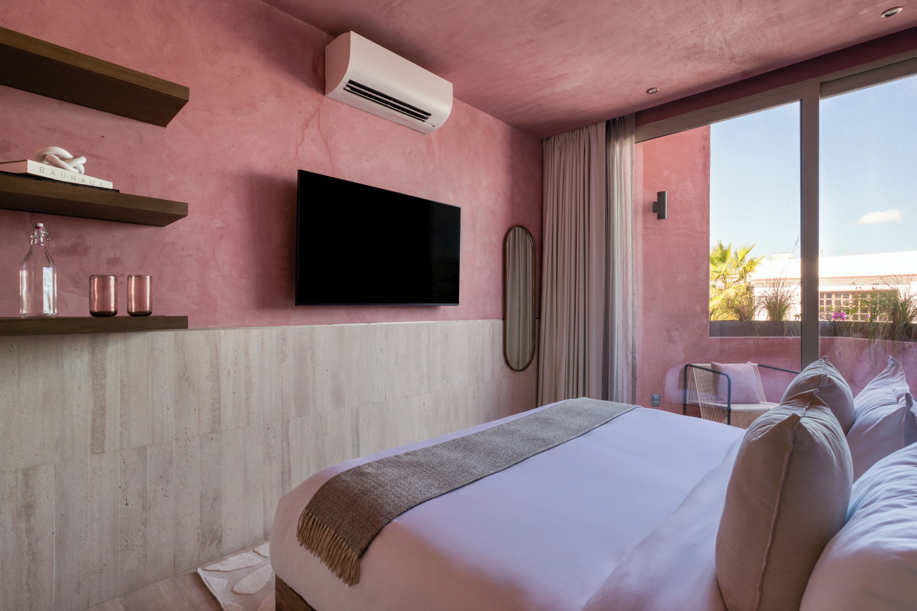 A hotel room with a large bed, a flat-screen TV mounted on the wall, a small table with a mirror, and a window with a view of palm trees.