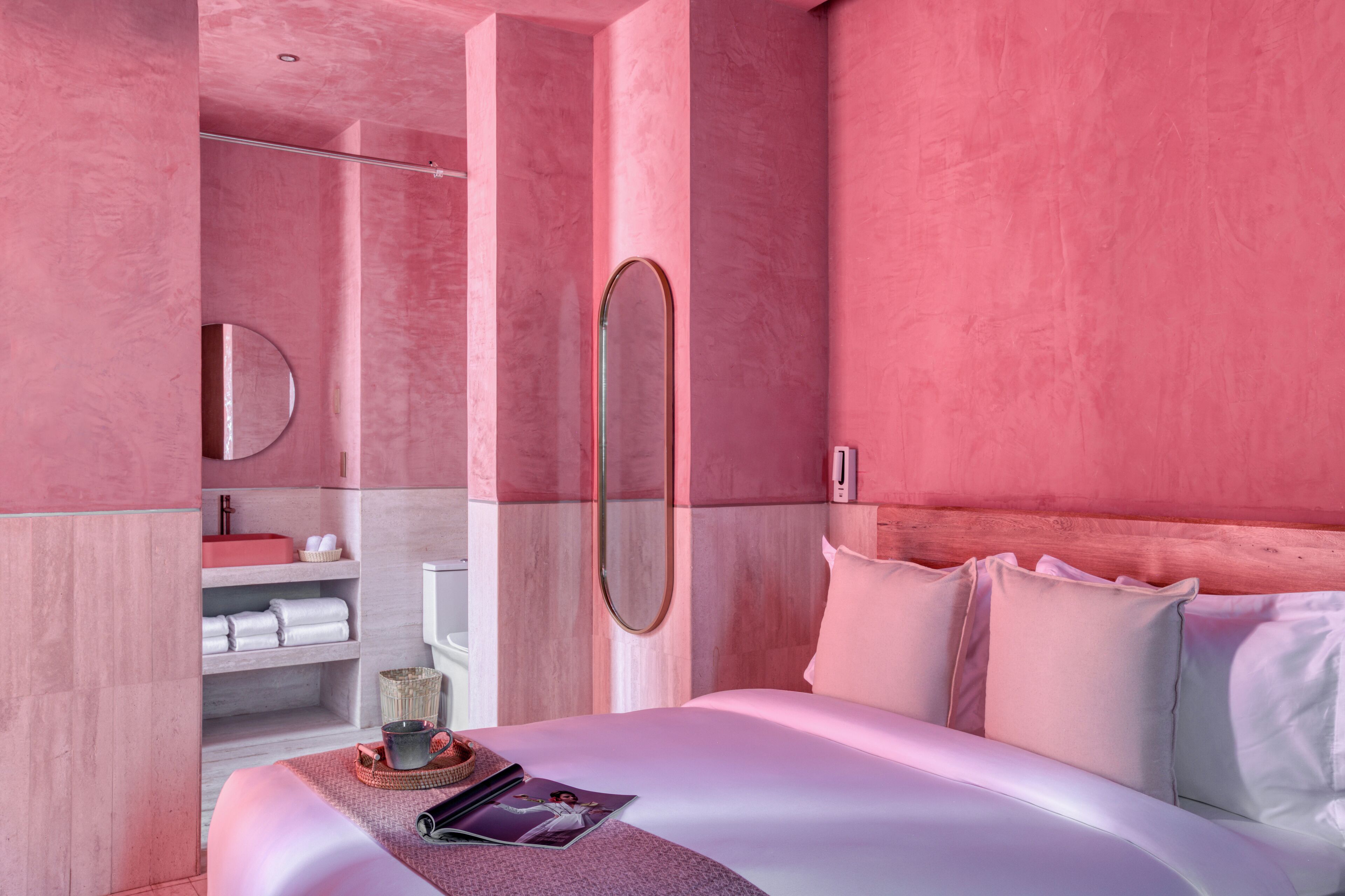 A modern bedroom with pink walls, a large bed, a bedside table with a magazine, and a bathroom with a toilet and towel rack.