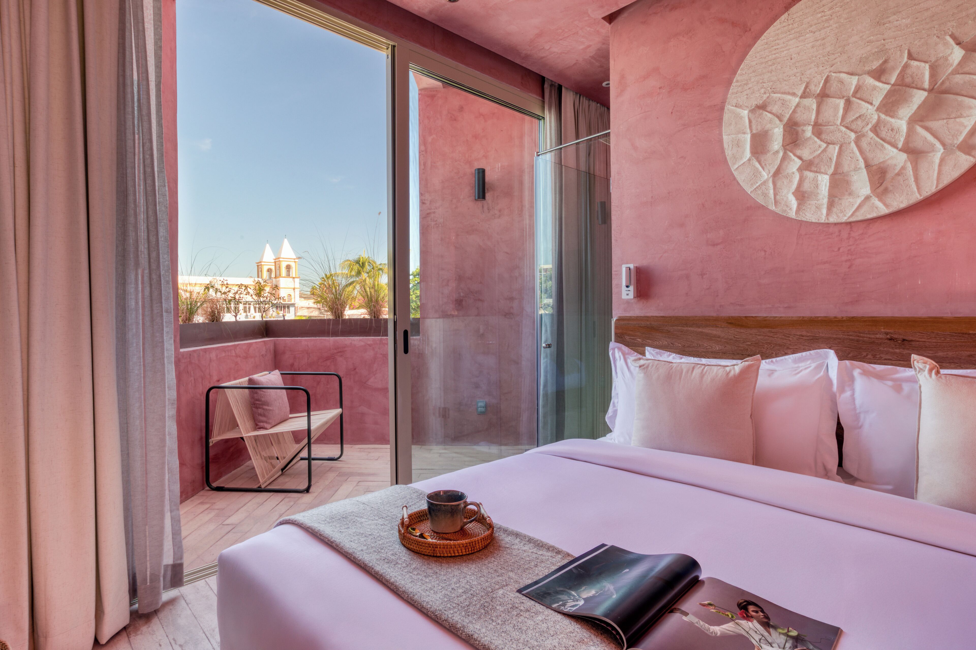 A hotel room with a large bed, a view of a building through a glass door, and a balcony with a wicker chair.