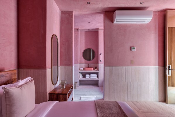 A pink-themed hotel room with a bed, a bedside table, a mirror, a small sink, and a towel rack.