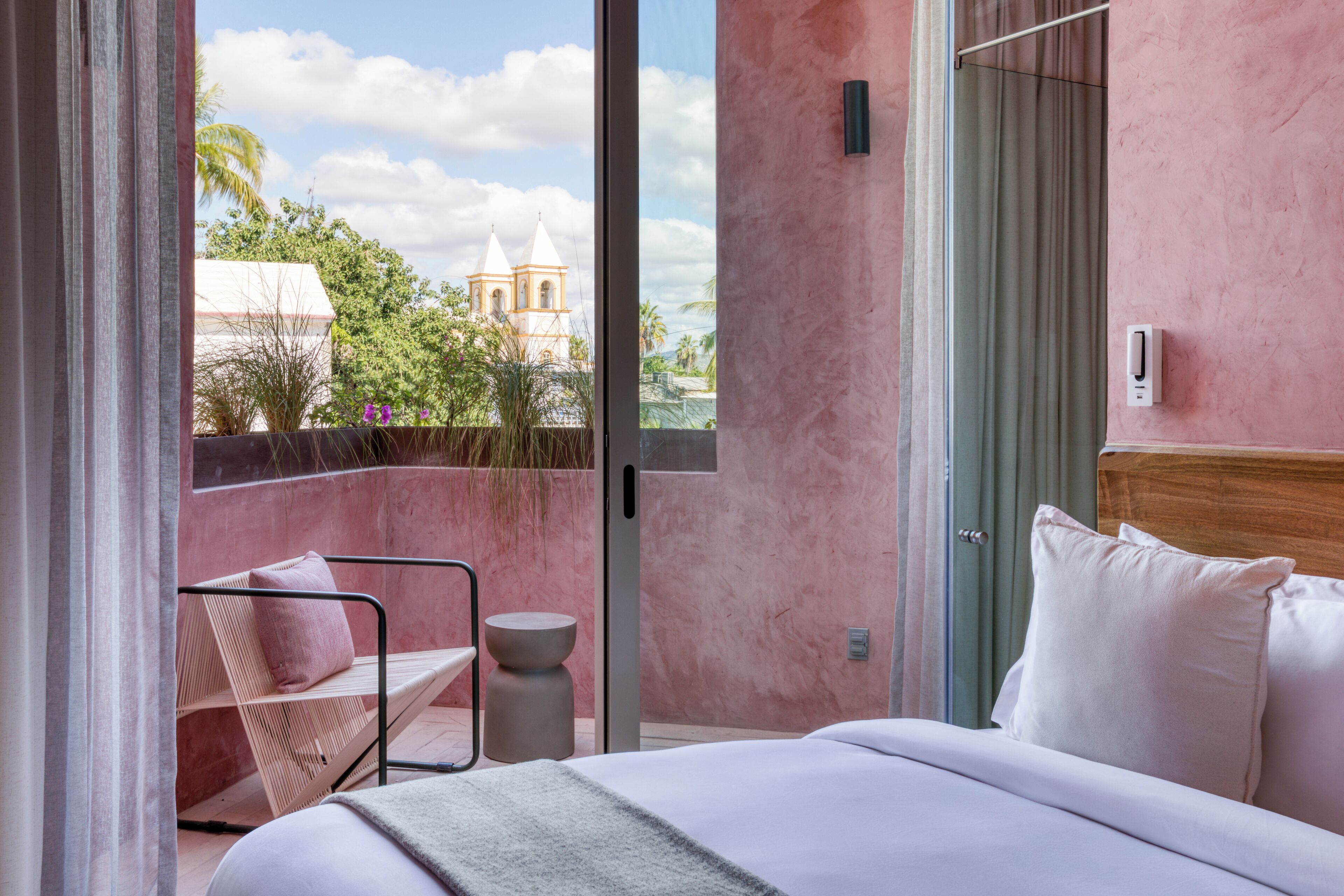 A hotel room with a balcony, a bed with white linens, a wooden headboard, and a view of a church and palm trees.