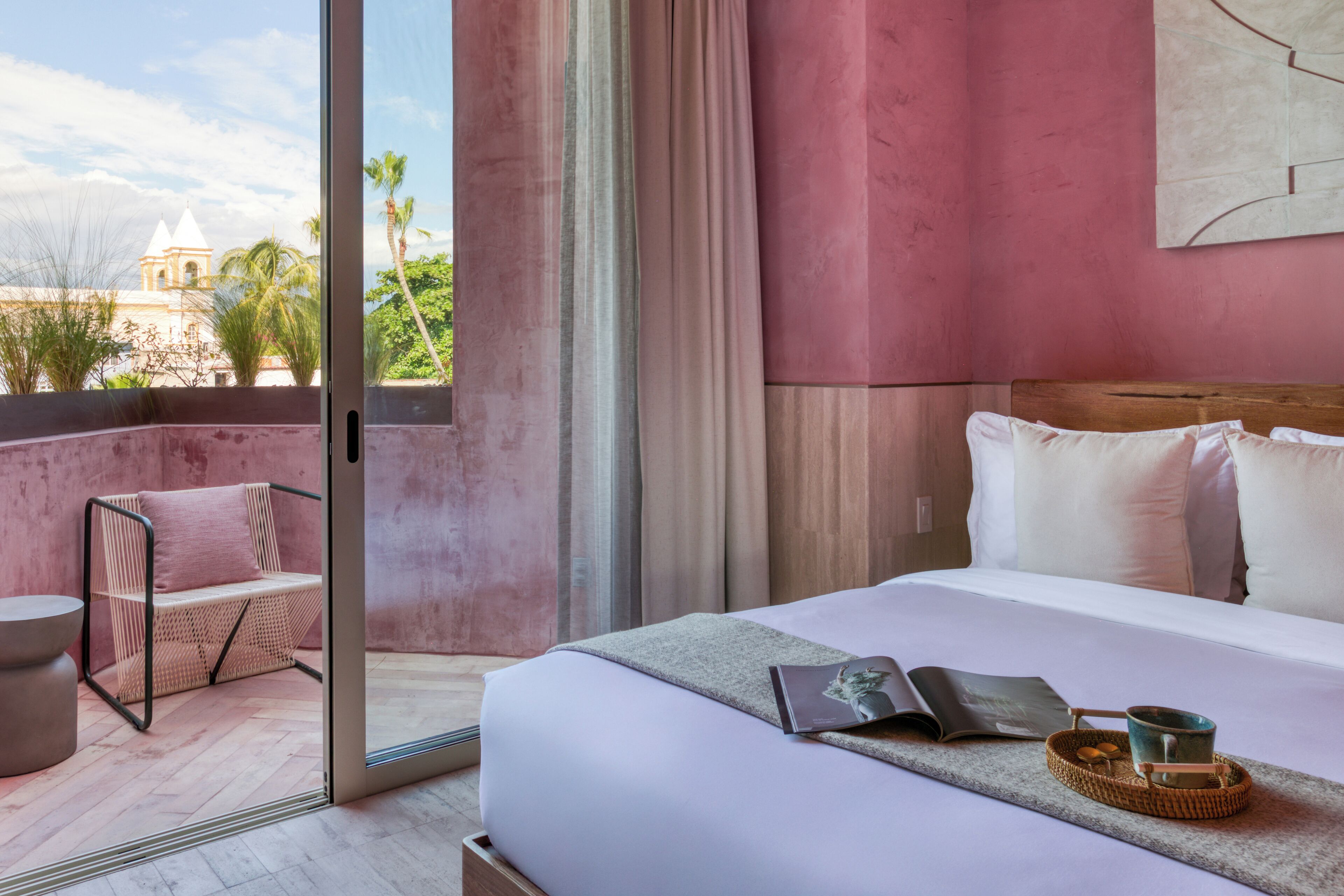 Ein Hotelzimmer mit Bett, Korbsessel, kleinem Tisch und Blick auf ein Gebäude.