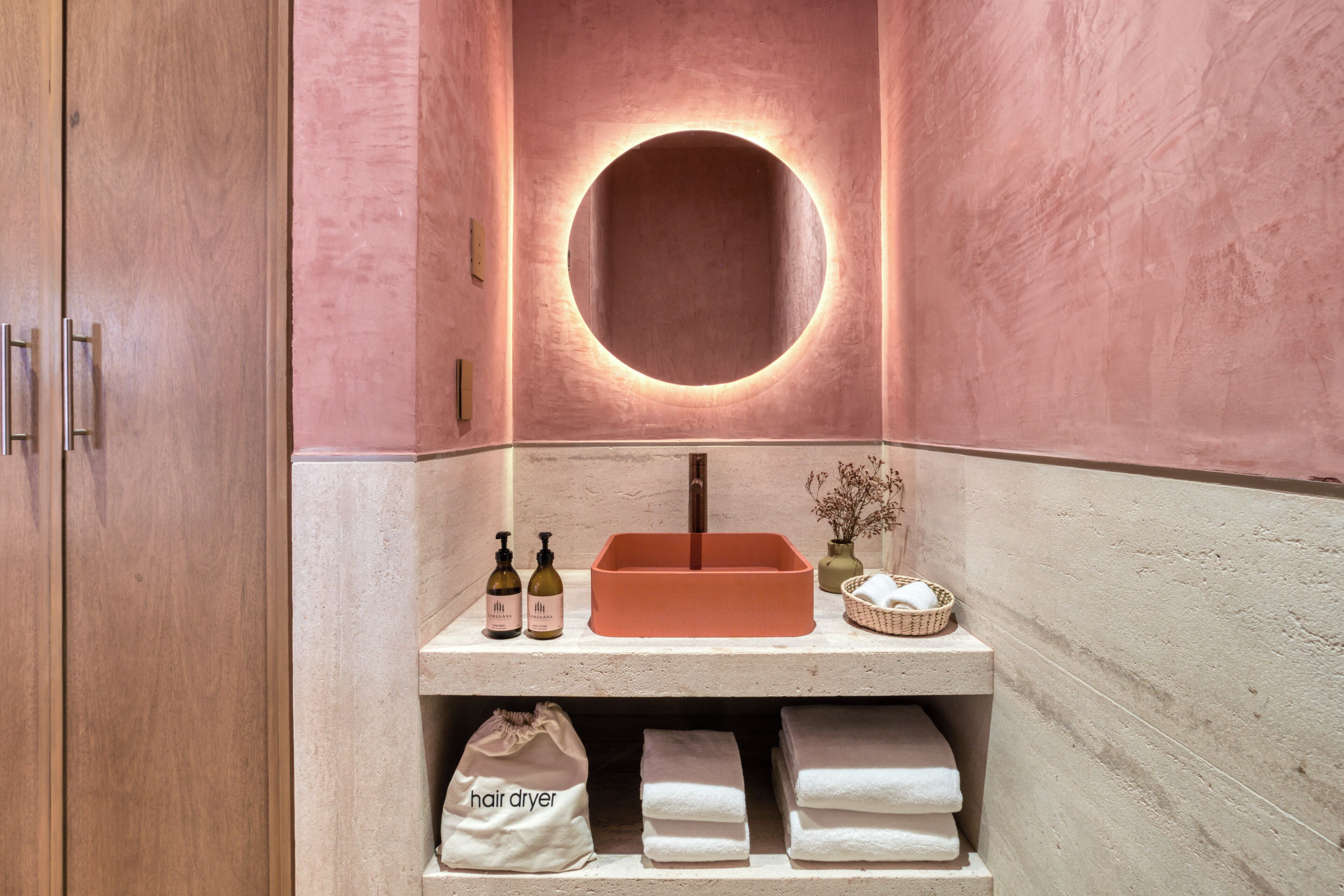 A bathroom with a wooden cabinet, a round mirror with a light, a shelf with toiletries, and a hair dryer bag.