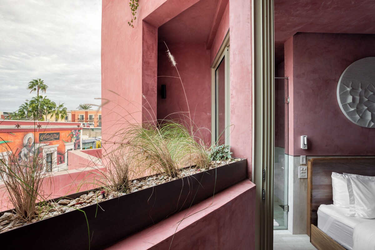 A hotel room with a bed, a window with a view of palm trees, and a decorative wall piece.