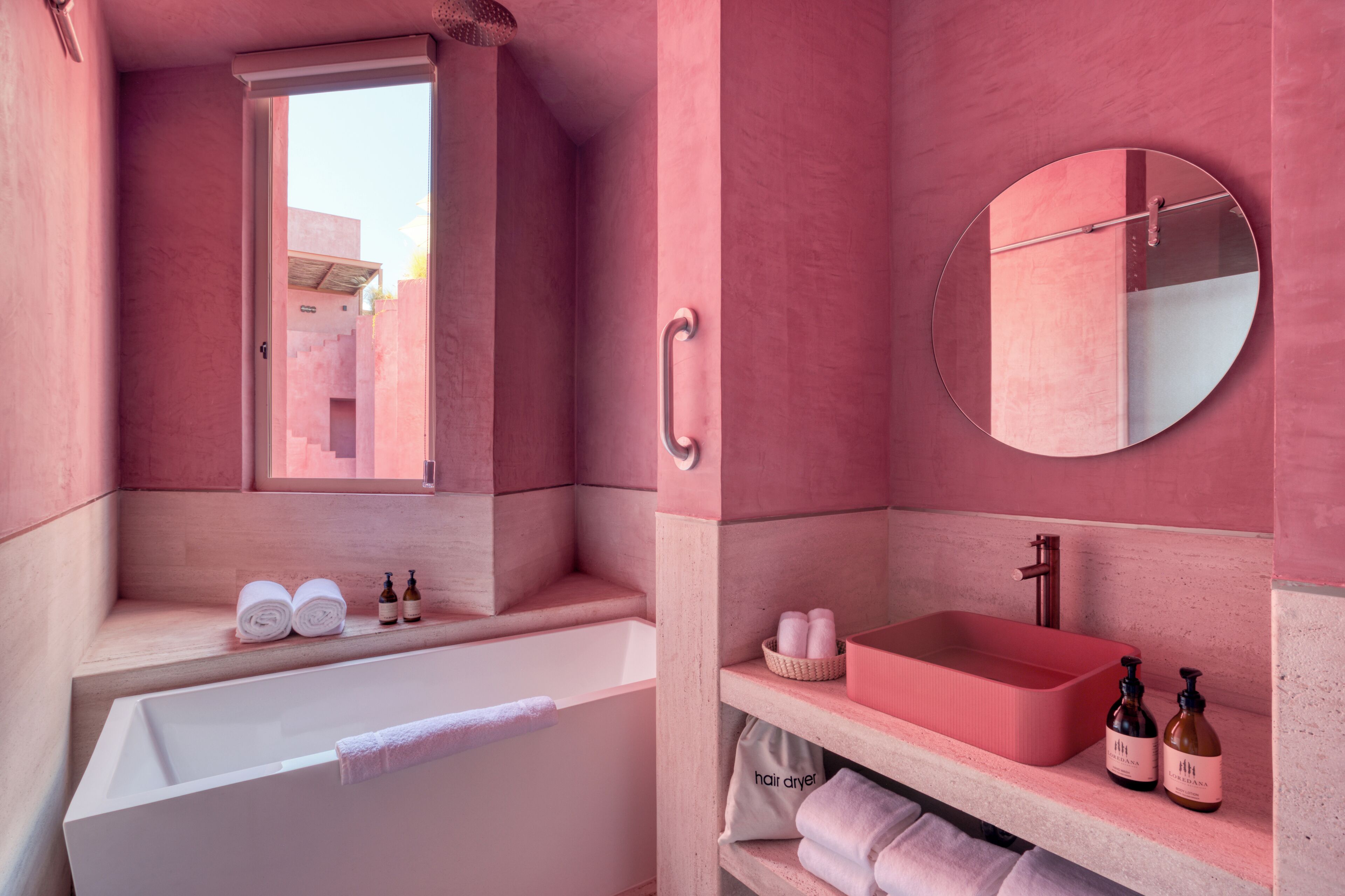 Un baño con decoración rosa, que incluye una bañera blanca, un espejo redondo, un lavabo con cuenca roja y un estante con toallas y artículos de aseo.