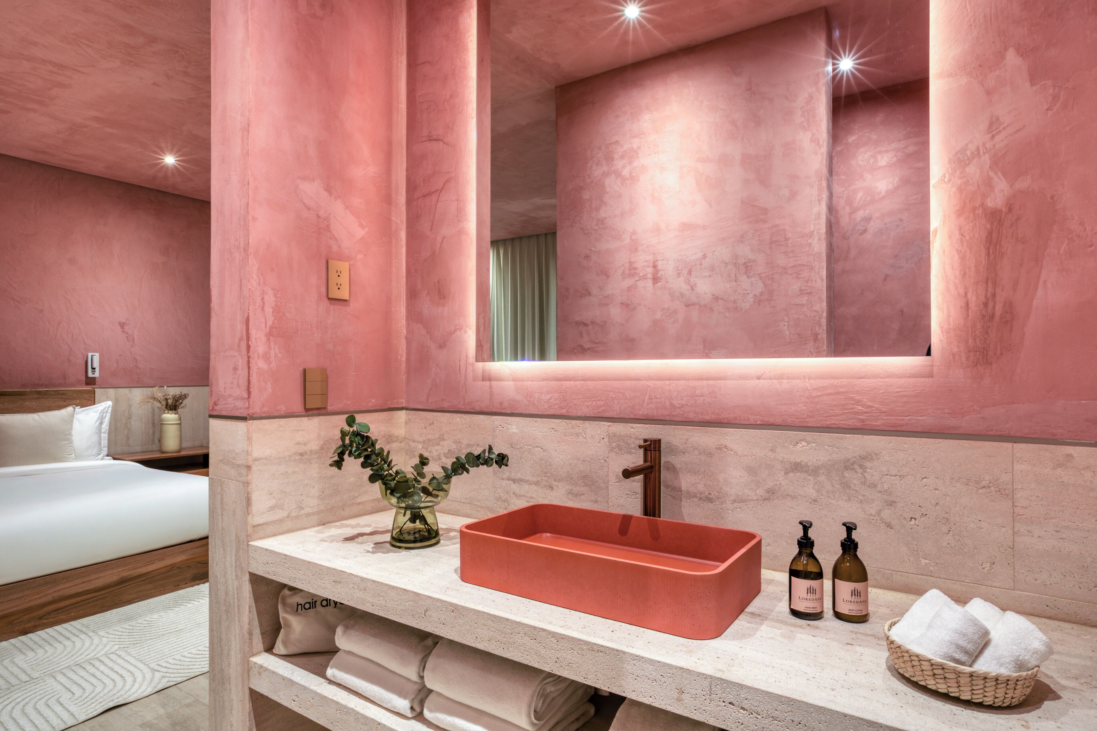 Een moderne badkamer met een roze wand, een rechthoekig wastafelmeubel en een spiegel erboven. Handdoeken en toiletartikelen liggen op het aanrecht.
