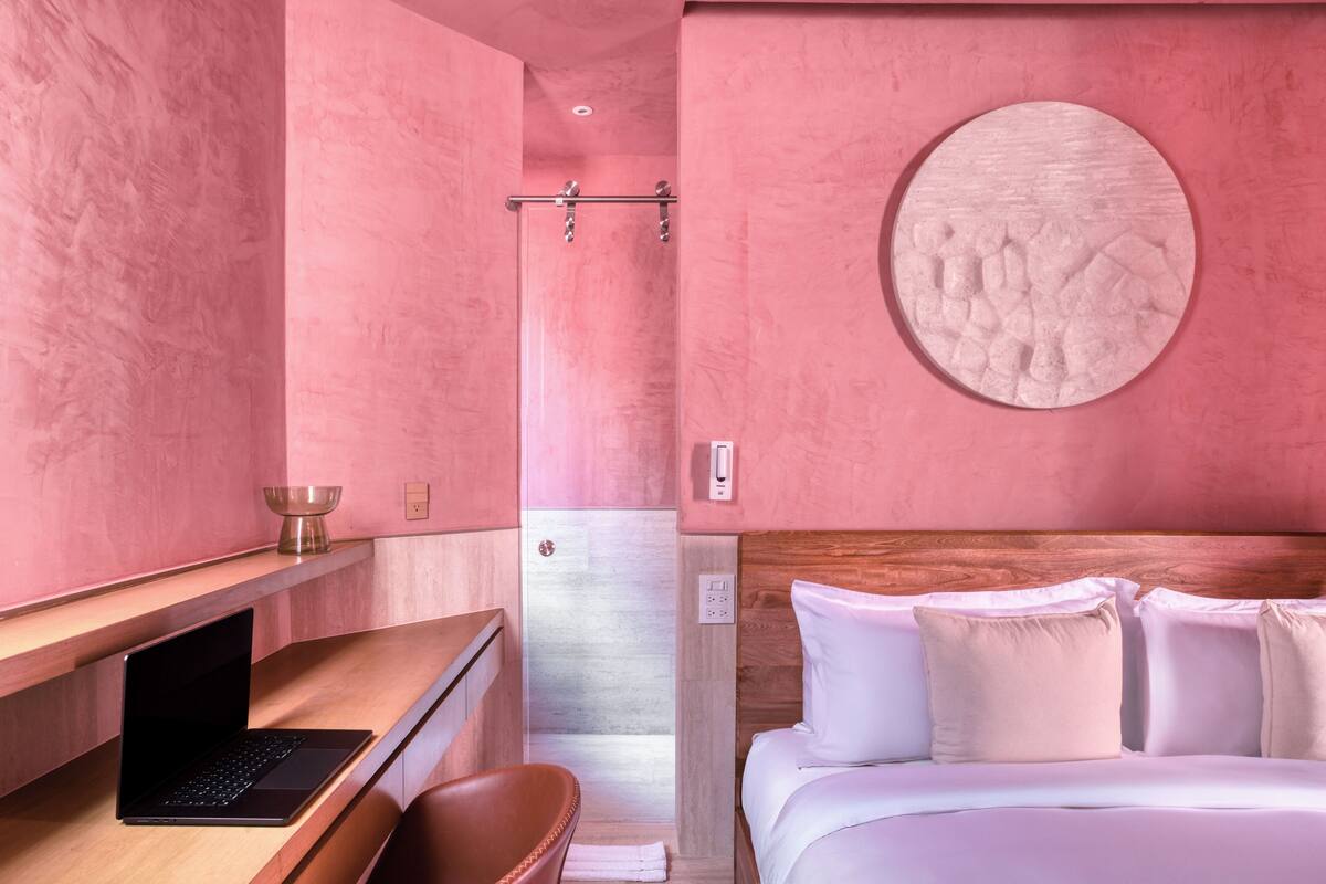 Een hotelkamer in roze tinten met een houten bureau, een laptop, een bed met witte beddengoed en een ronde wanddecoratie.