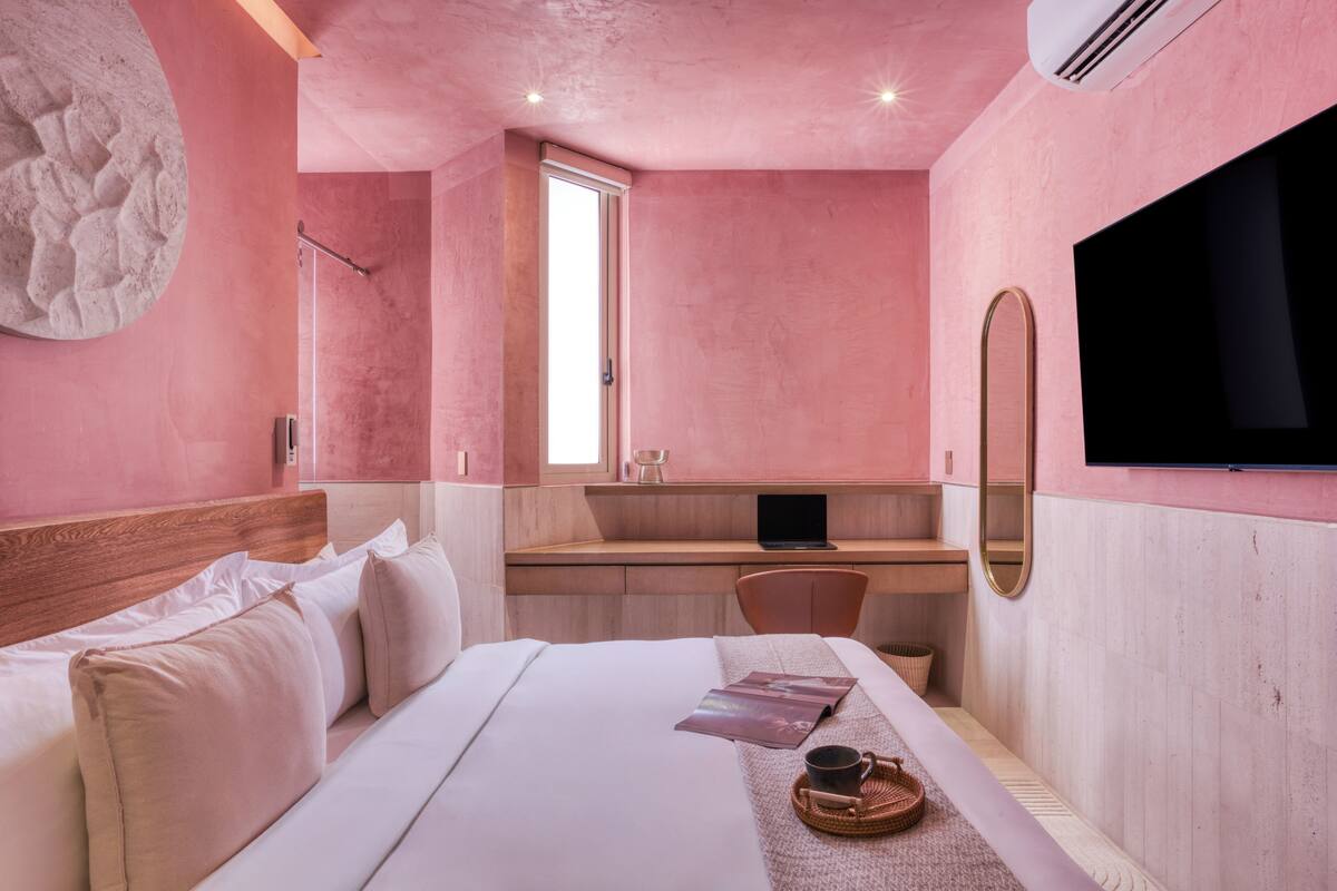 Een hotelkamer in roze tinten met een bed, een bureau, een stoel, een televisie en een grote spiegel.