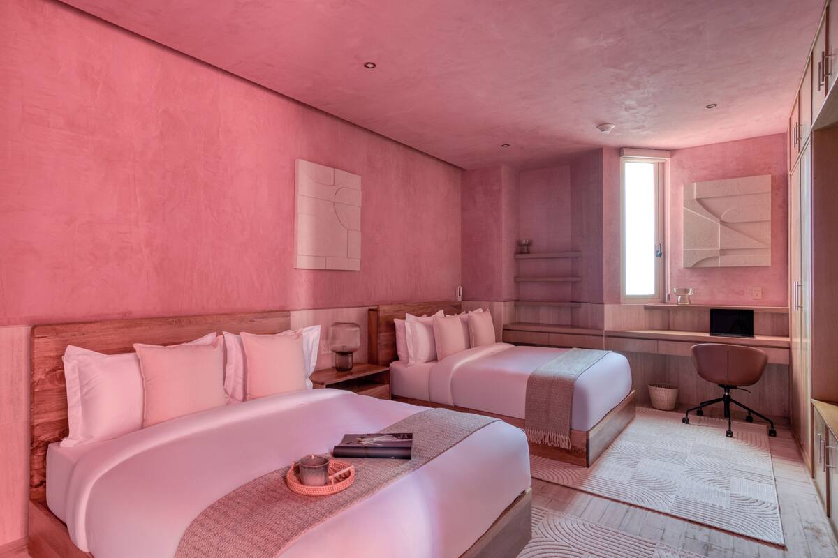 Een hotelkamer in roze tinten met twee bedden, een bureau, een stoel en een grote spiegel.