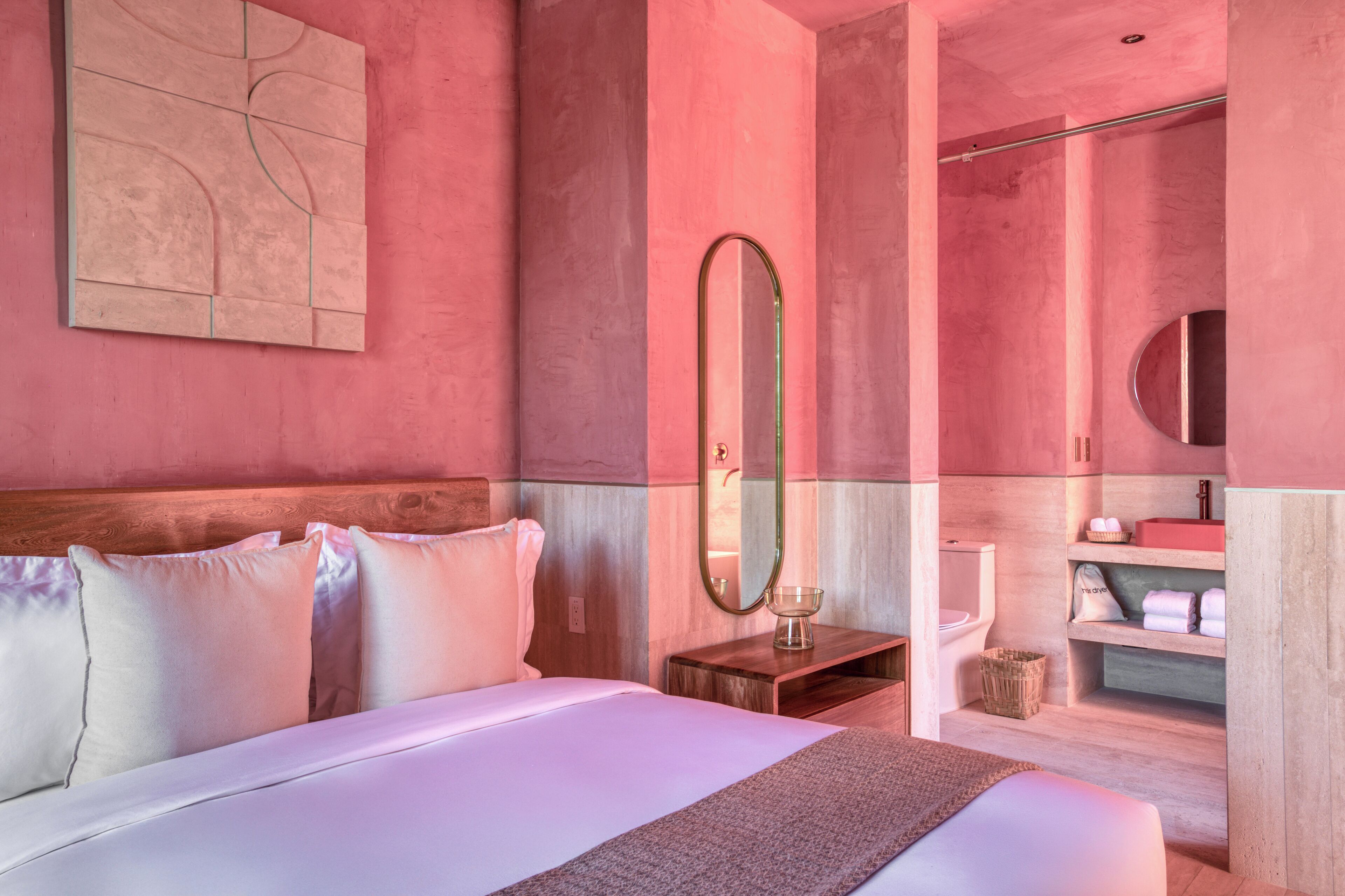 A hotel room with a large bed, a wooden headboard, a bedside table with a mirror, a small wooden stool, and a bathroom with a toilet, sink, and shelves.