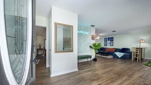 Una sala de estar con piso de madera, un banco verde, un sofá con cojines naranjas, un espejo grande y un letrero de neón en la pared.