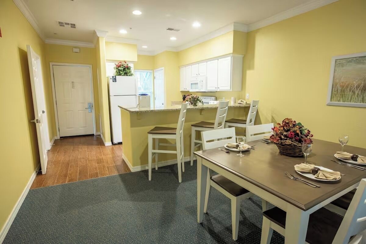 A dining area with a table set for two, chairs, a refrigerator, and a microwave in the background.