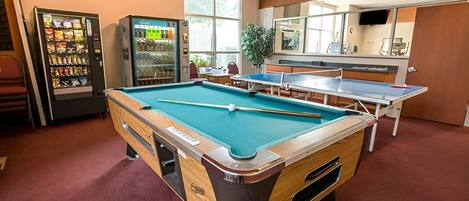 A pool table in a room with a vending machine and a ping pong table.