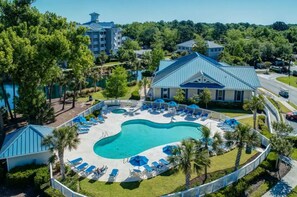 An aerial view of a resort with a large swimming pool surrounded by lounge chairs and umbrellas, a golf course, and a mix of residential and commercial buildings.