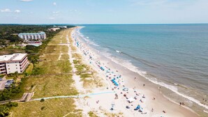 An aerial view of a beach with a long stretch of sand, a few buildings, and a clear sky.