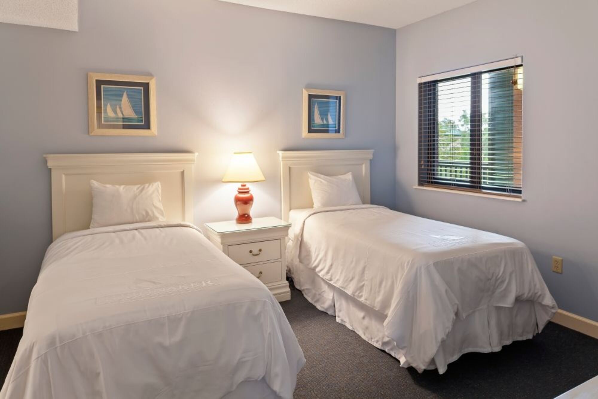 A bedroom with two single beds, a nightstand with a lamp, and two framed pictures on the wall.