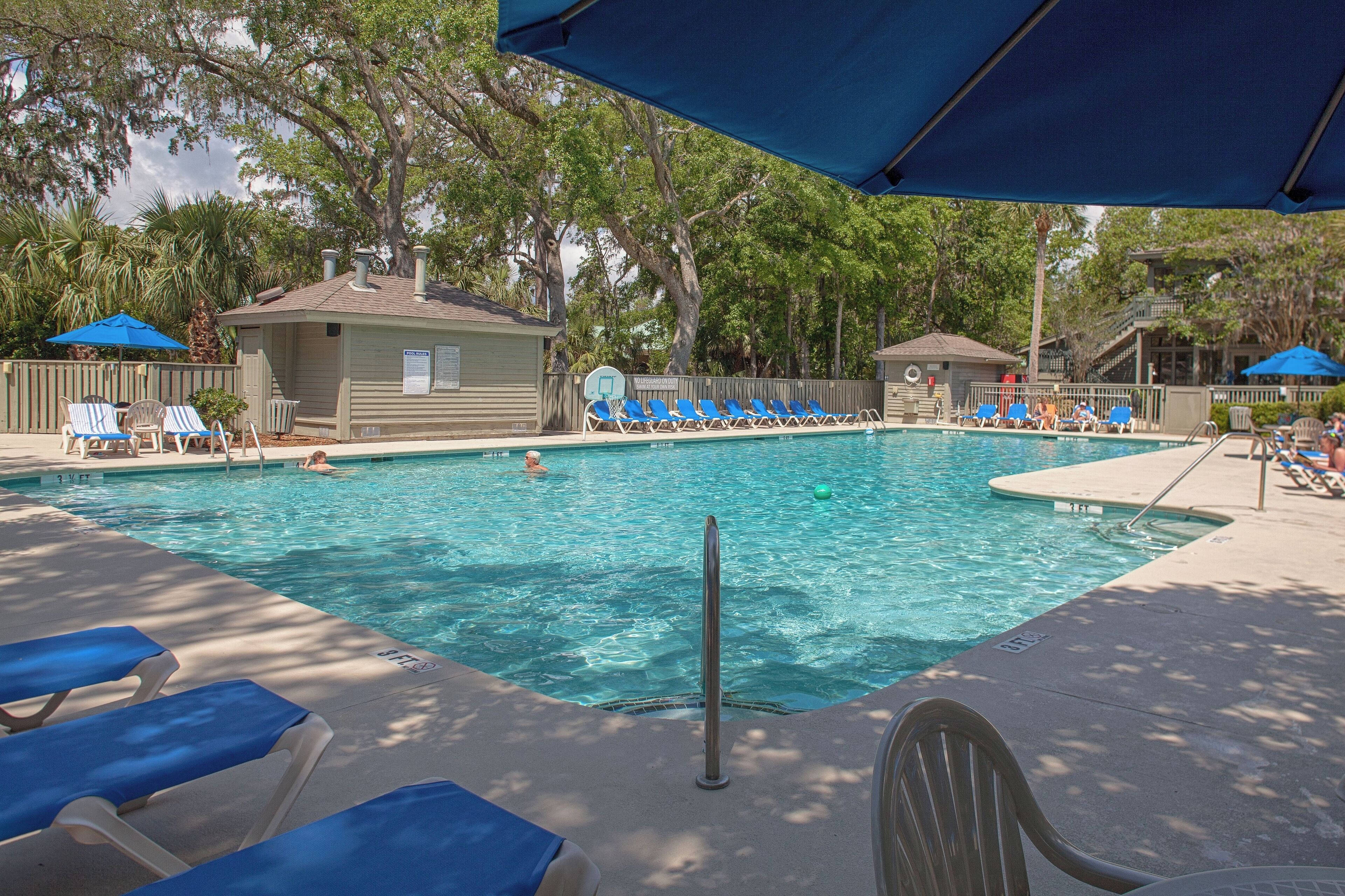 A swimming pool with blue lounge chairs and umbrellas, surrounded by trees and a small building.