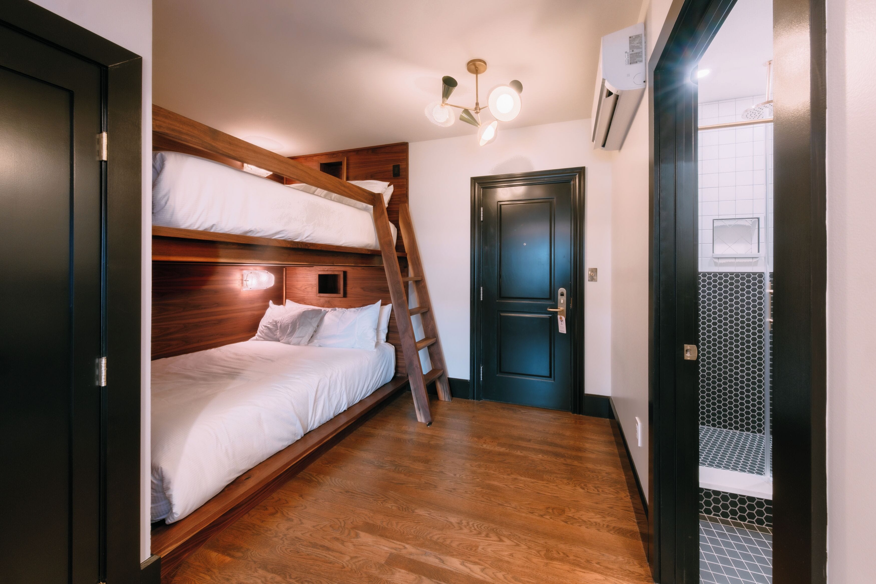 Una habitación con literas, una escalera de madera, una puerta oscura y una lámpara en el techo.