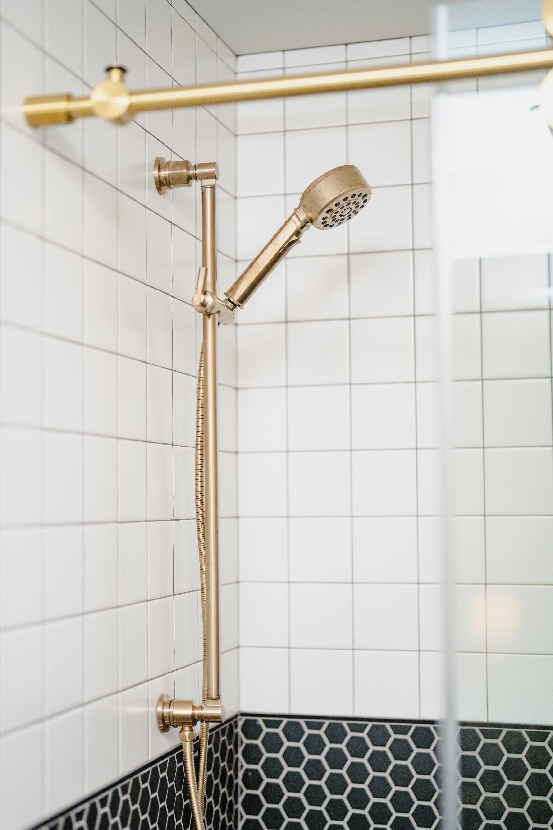 Eine Dusche mit goldfarbenem Duschkopf und Duschschlauch, weißen Fliesenwänden und einem schwarz-weißen Muster im unteren Bereich.