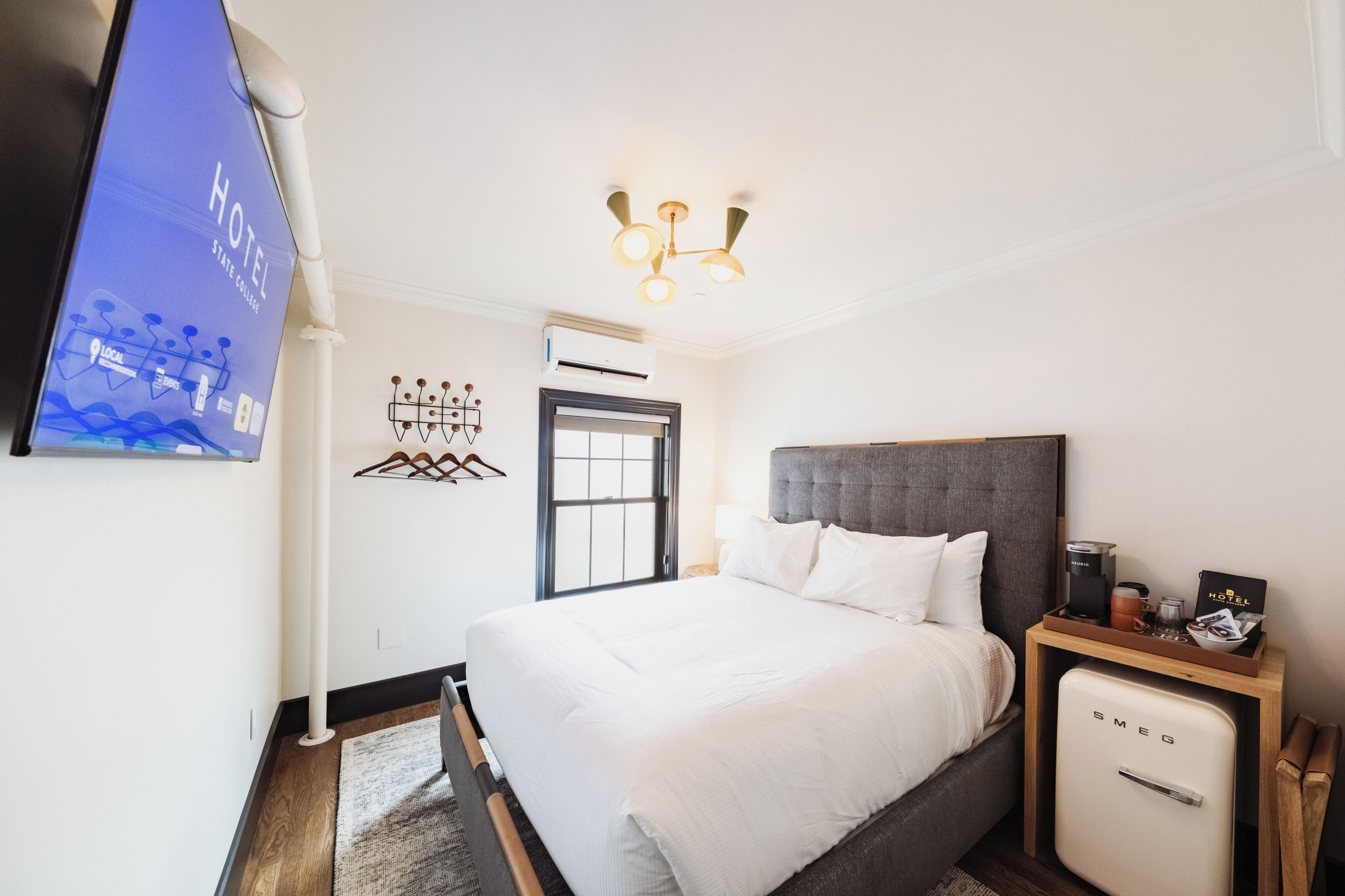 A hotel room with a bed, a nightstand with a coffee maker, a minibar, and a wall-mounted TV displaying the hotel's name.