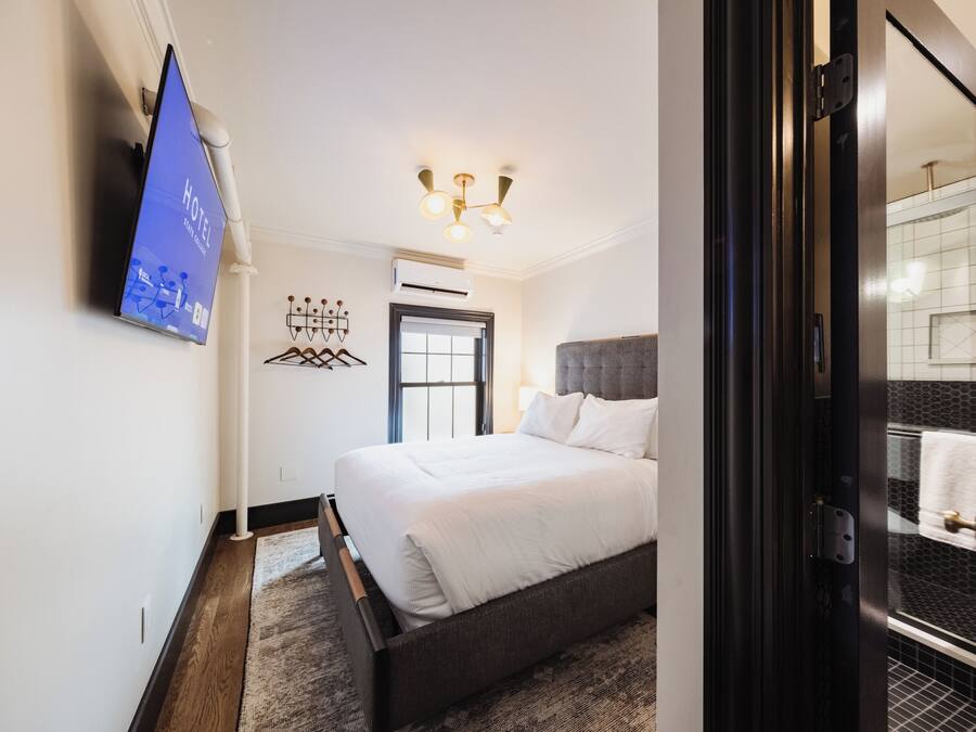 Een moderne hotelkamer met een bed, een aan de muur bevestigde televisie, een plafondlamp en uitzicht op de badkamer door een glazen deur.