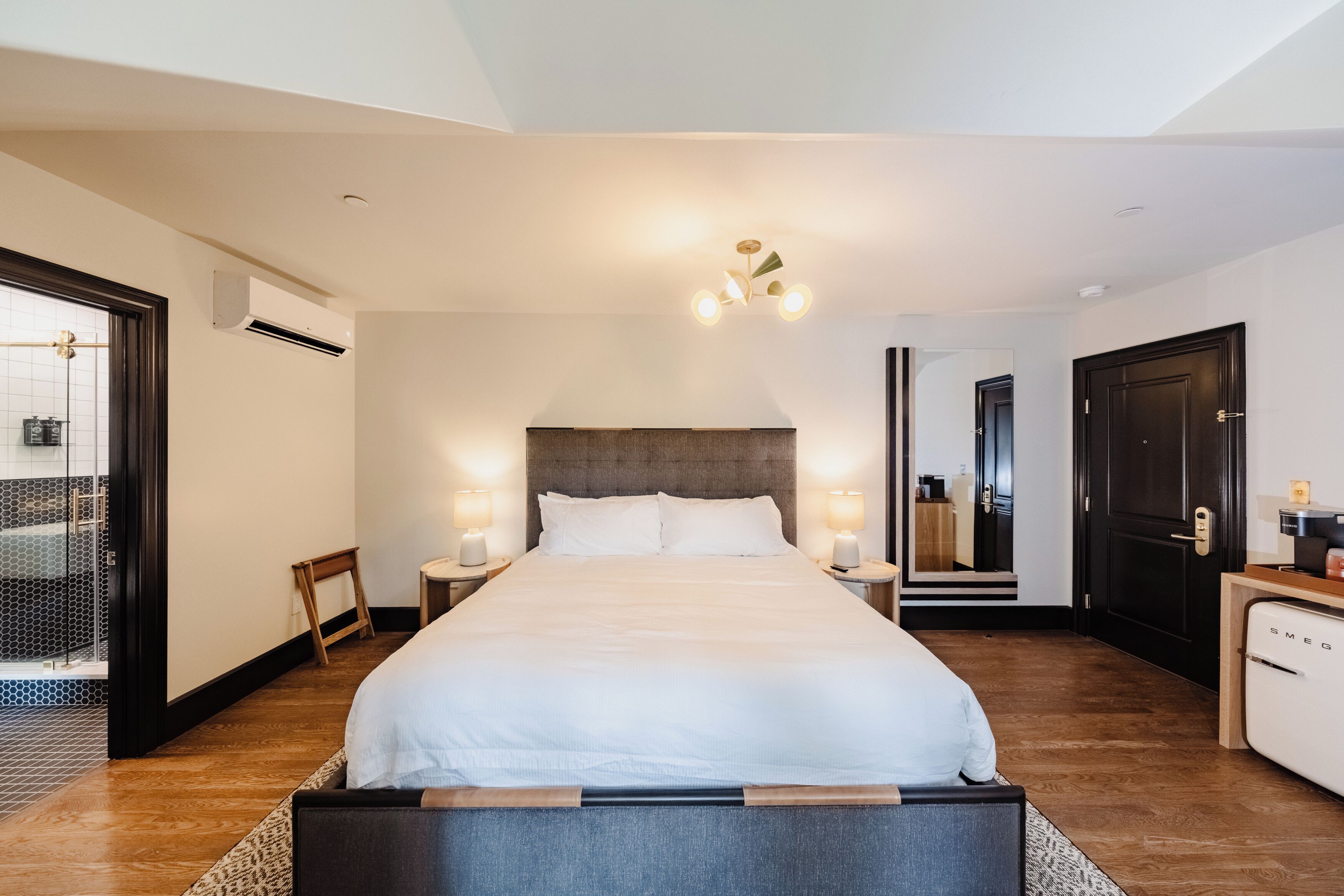 Una habitación de hotel moderna con una cama grande, lámparas de noche, un banco de madera, una mesita y un refrigerador integrado.