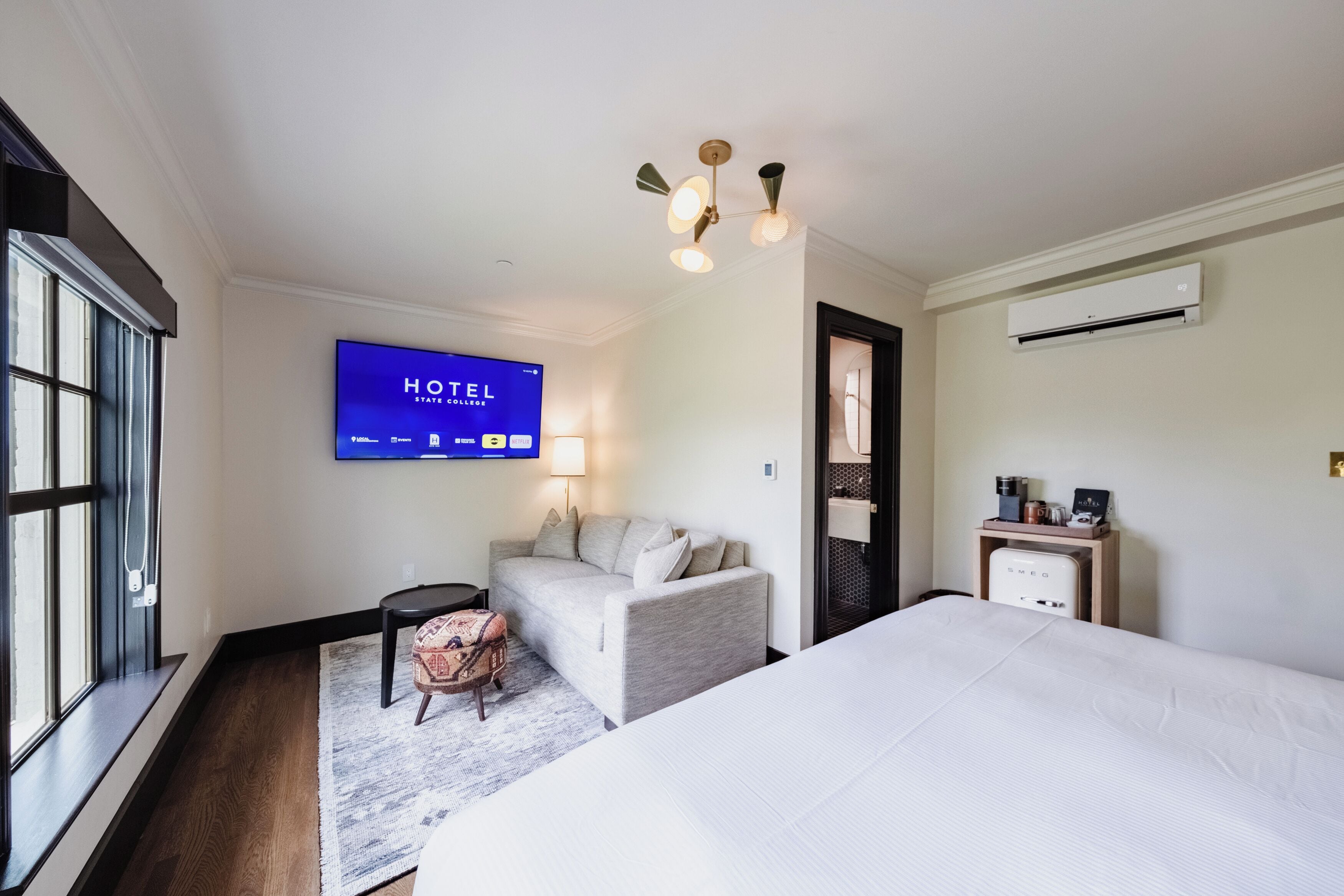 A hotel room with a bed, a sofa, a TV displaying the hotel's name, a lamp, and an air conditioning unit.