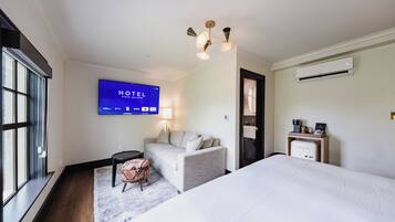 A hotel room with a bed, a sofa, a TV displaying the hotel's name, a lamp, and an air conditioning unit.