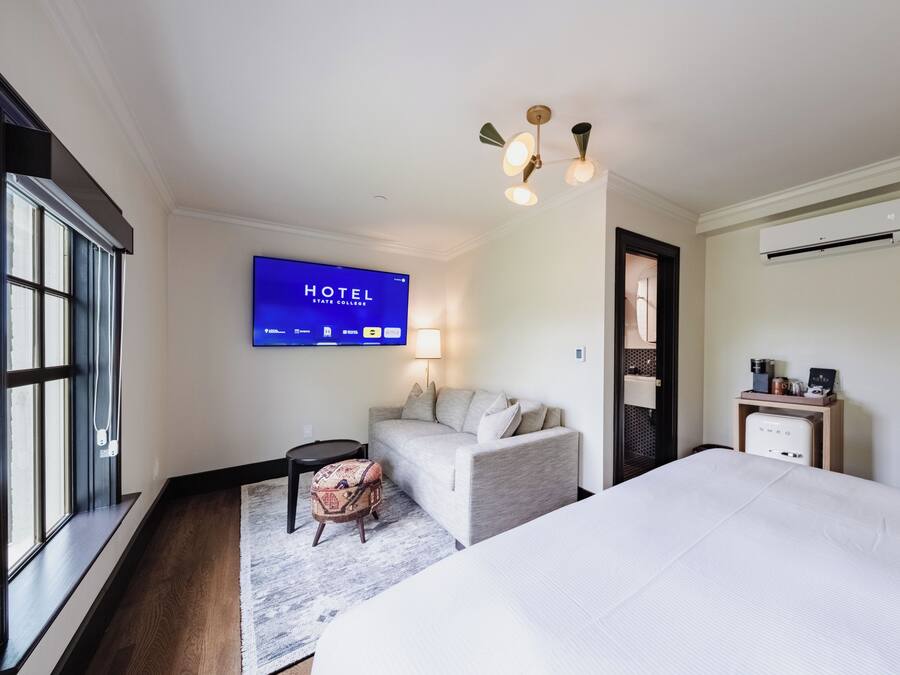 A hotel room with a bed, a sofa, a TV displaying the hotel's name, a lamp, and an air conditioning unit.