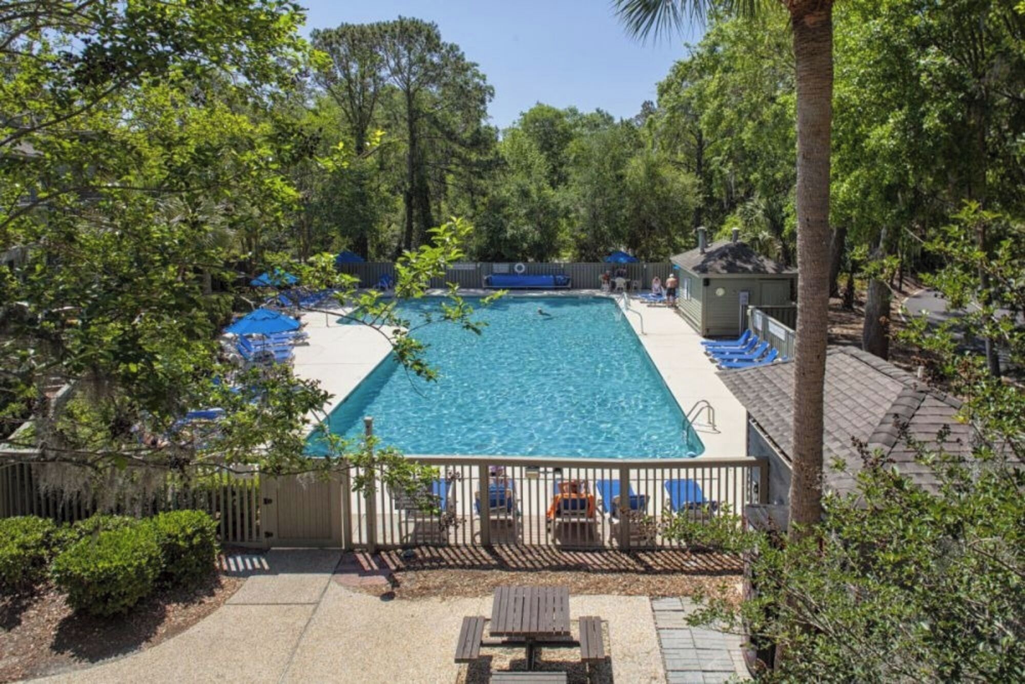 A rectangular pool surrounded by lounge chairs and umbrellas, with a wooden deck and a small table in the foreground.