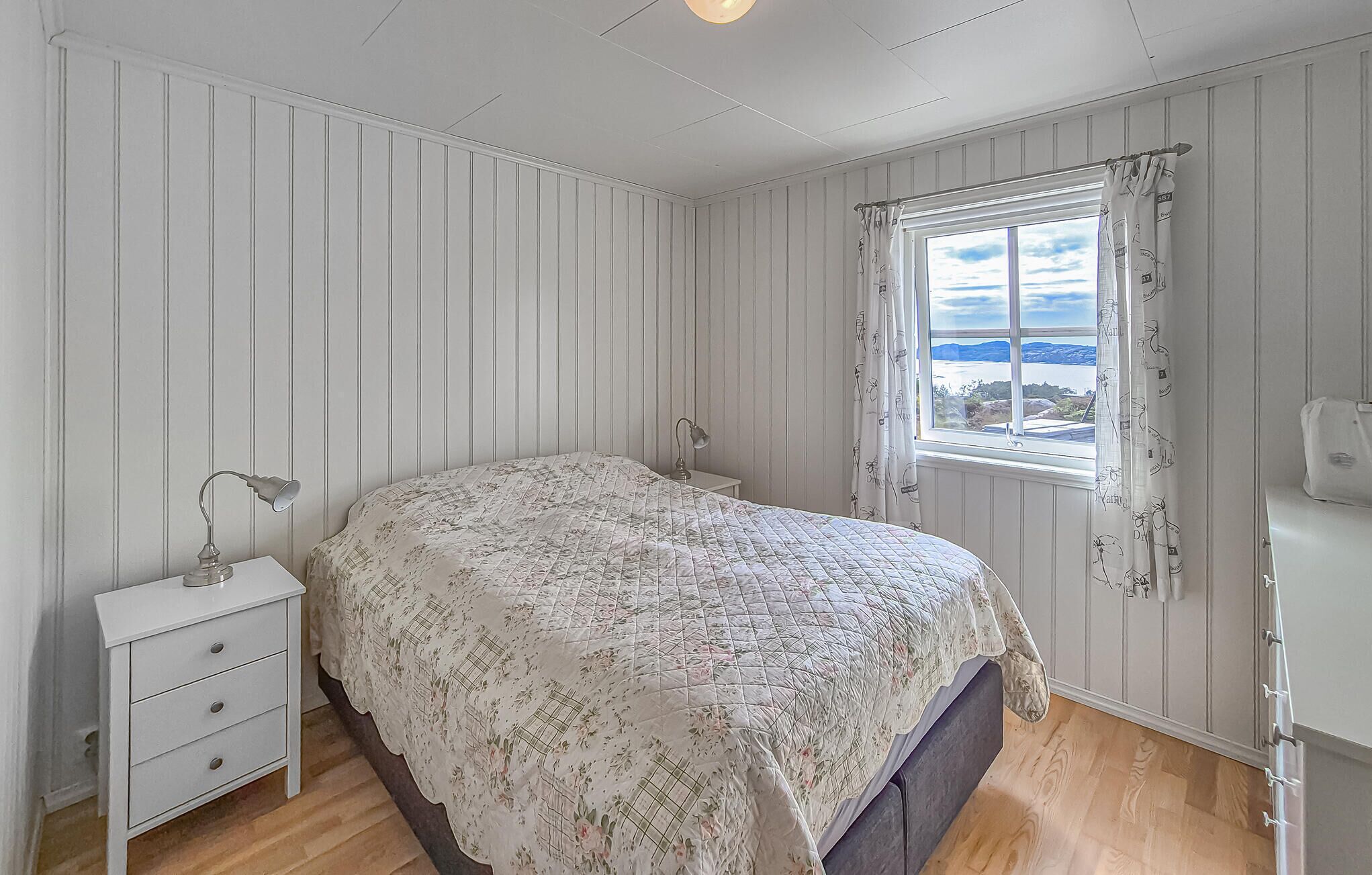 Un dormitorio con cama, mesita de noche, lámpara, ventana con cortinas y vistas al exterior.