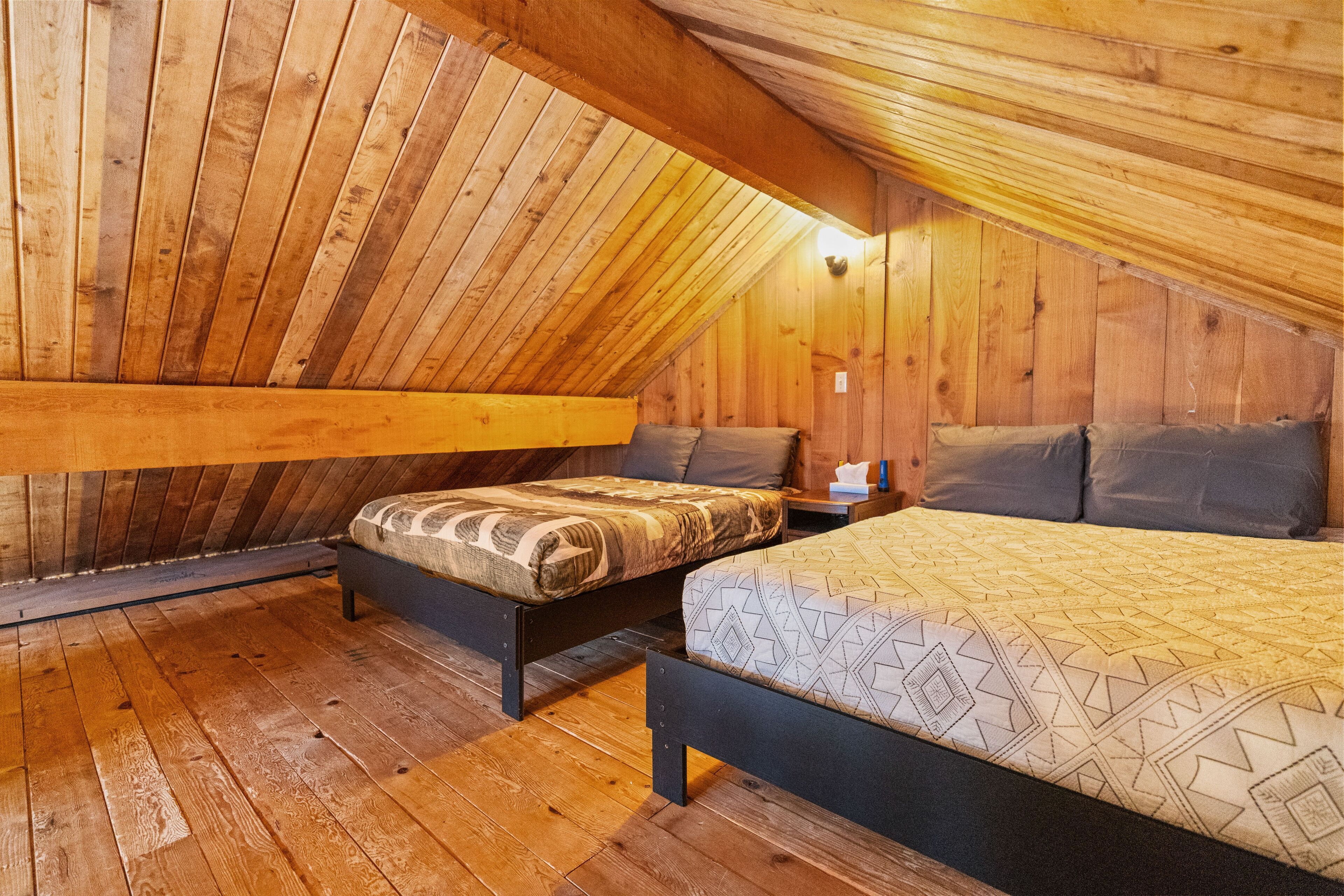 Une chambre mansardée en bois avec deux lits, une table de chevet et une lampe fixée au mur.