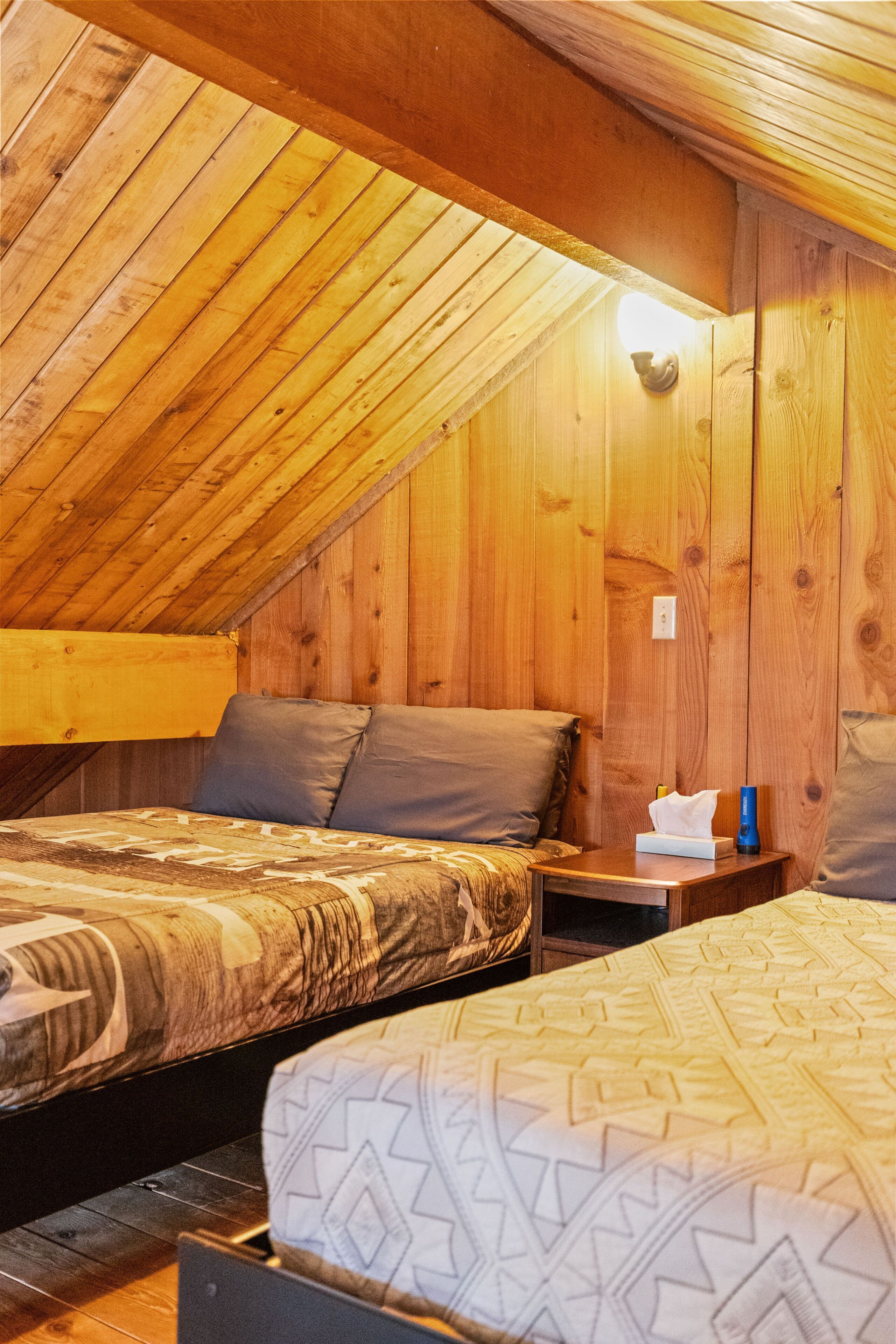 Un acogedor dormitorio en el ático con paredes de madera y techos inclinados. Hay dos camas con ropa de cama estampada y una mesita de noche pequeña con una lámpara.