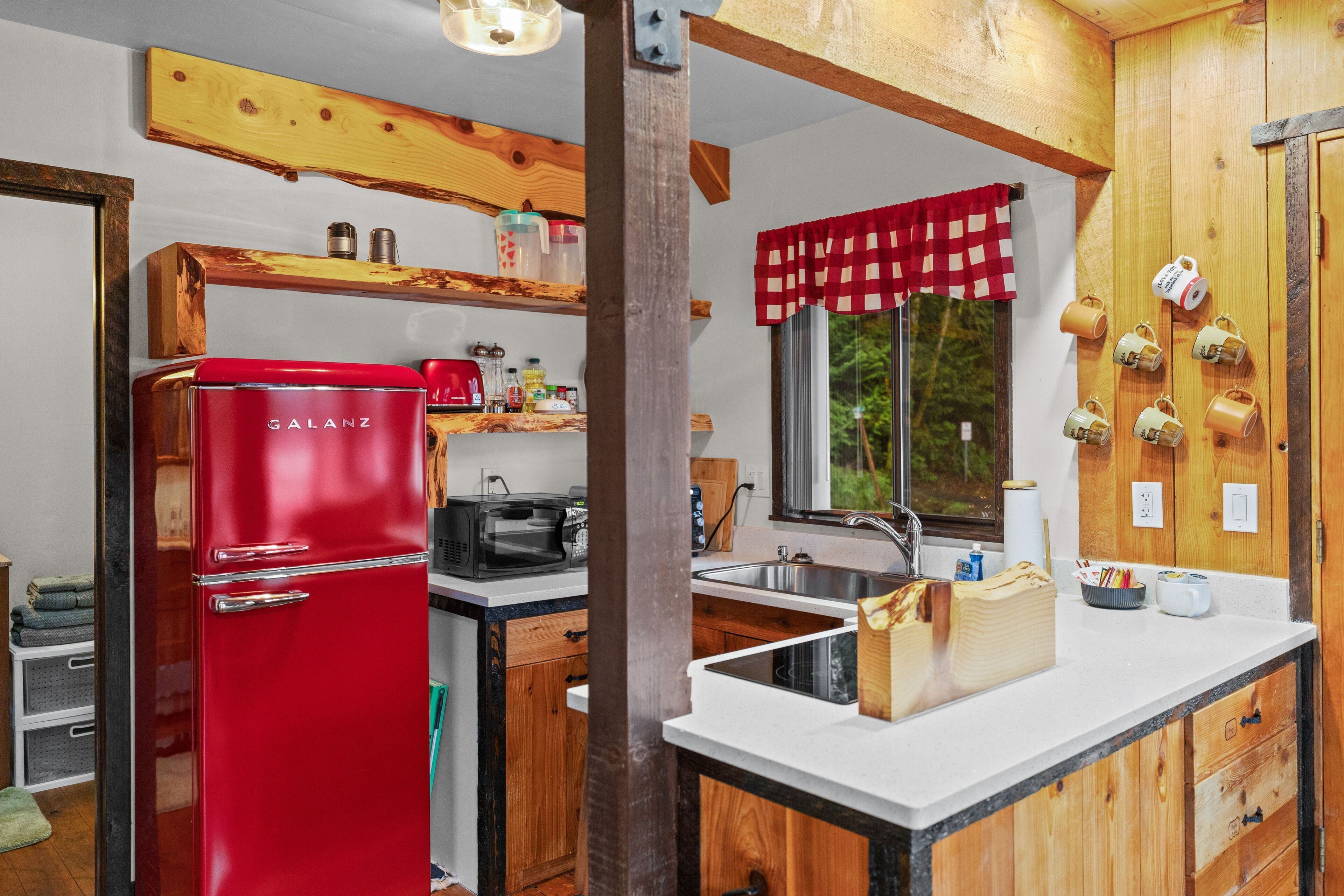 A compact kitchen with wooden cabinetry, a red Galanz refrigerator, a coffee machine, and a window with a checkered curtain.