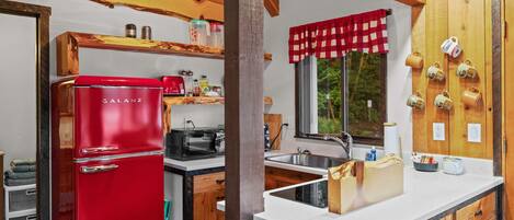 A compact kitchen with wooden cabinetry, a red Galanz refrigerator, a coffee machine, and a window with a checkered curtain.