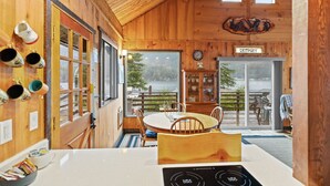 A wooden interior with a dining area, a kitchen with an induction cooktop, and a view of the outdoors. - Lake Sutherland Cabin w/ Dock Access & Kayaks (Port Angeles)